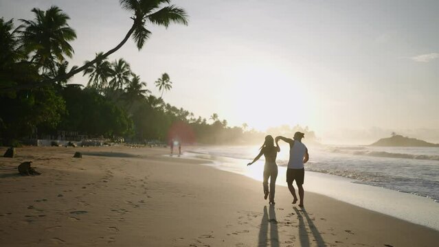 Young happy couple holding hands and running and fooling around on the beach together enjoying summer back view. Boyfriend and girlfriend having fun at the seaside chasing each other at sunrise.