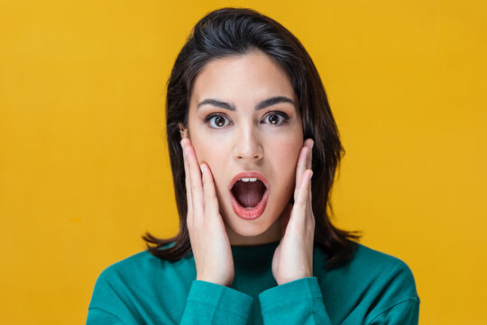 Surprised young woman screaming and putting her hands on her face looking at camera isolated on yellow