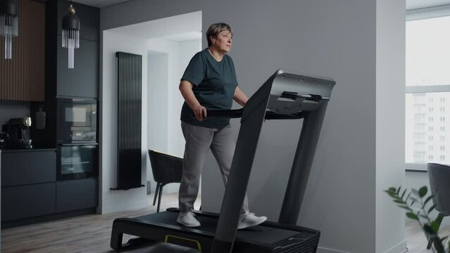 Mature Woman Walking On Treadmill In Living Room of Modern Apartment, Healthy Lifestyle In Old Age