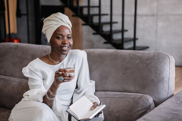 Portrait of satisfied African young woman in white turban sitting on couch holding glass of water looks aside thoughtfully at home. Purposeful Brazilian businesswoman in traditional african clothes.