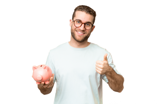Young man holding a piggybank over isolated chroma key background with thumbs up because something good has happened