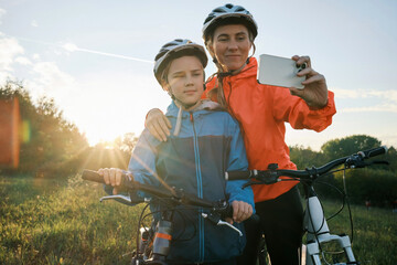 Mother and son ride a bike. Happy cute boy in helmet learn to riding a bike in park on green meadow in autumn day at sunset time. Family weekend.

