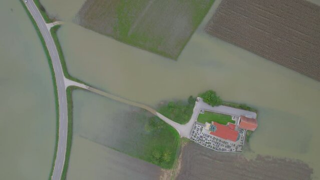 AERIAL TOP DOWN: Church with cemetery is inaccessible due to flooded access road. Village church on a dry island surrounded by muddy flood water and submerged farmland after abundant autumn rainfall.