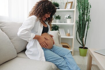 Pregnant woman smile and happiness sits on the couch freedom and strokes her belly feels kicks with the baby in the last month of pregnancy, mother's day lifestyle