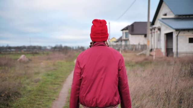 A sad teenage girl in red clothes is walking down the street. View from the back.