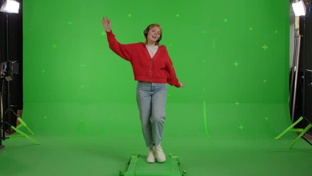 Attractive red-haired white girl walks to the side against the background of a green screen. Happy young woman in jeans and a knitted jumper listens to music on headphones and dances.