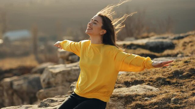 A young woman in a yellow sweater sits on the edge of a rock and raises her arms to the sides imitating flight, happy and cheerful admiring nature, wind blowing her hair in the sunlight, traveling