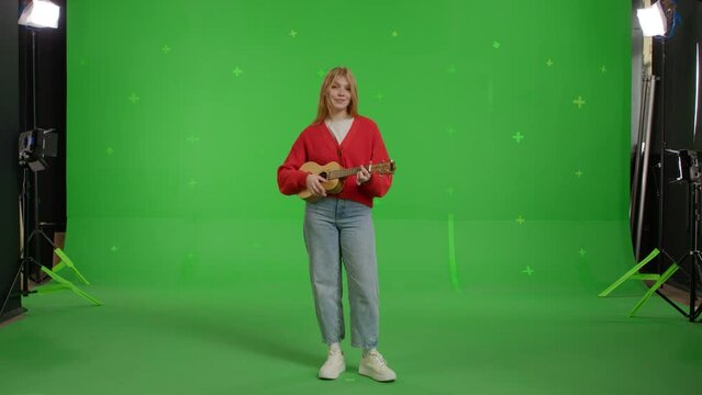 Attractive red-haired white girl plays the ukulele against the background of a green screen. Happy young woman in jeans and knitted jumper enjoying music
