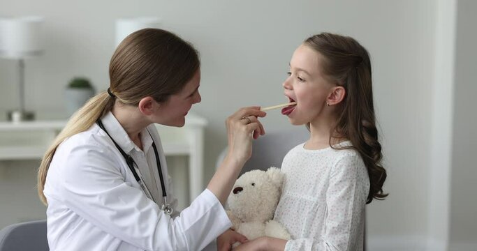 Focused pediatrician examining kid in doctor office, checking sore throat, asking girl to open mouth, showing tongue, talking to preschool child with toy, visiting sick little patient at home