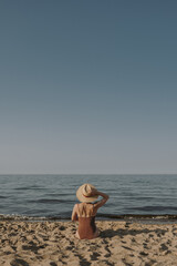 Young woman sitting and relaxing on the beach sand with view on sea. Harmony, lounge, summer vacation concept. Elegant fashion composition with straw hat and bikini swimsuit