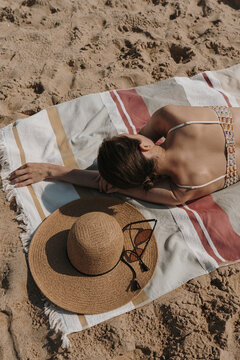 Beautiful woman chilling and sunbathing on the beach. Aesthetic stylish female sunglasses, straw hat, beach towel on beach sand. Elegant summer fashion concept