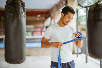 a asian male boxer wearing the boxing wrap gloves while standing with the box ring at the background