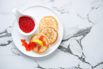 White plate with red caviar and mini pancakes, above view on a white marble background, horizontal...