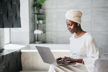 Purposeful African woman in white turban works at home using laptop looks screen. Confident Brazilian businesswoman remote managing company. American Female Writer typing text on computer at hotel.