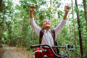 a male elementary student in uniform riding his bike through the country road and raised his hands in the air with a lot of tree at the background