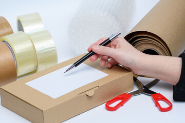 Karton z przesyłką i etykietą na tle akcesoriów do pakowania paczek takich jak folia bąbelkowa, taśma, nożyczki  i dłoń z długopisem w sklepieinternetowym  - obrazy, fototapety, plakaty
