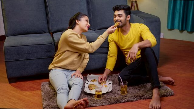 Happy young couple feeding each other with pizza while sitting on floor at home - concept of dating, affection and love relationship.