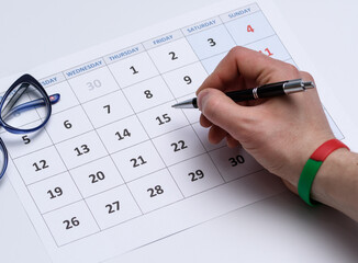 Kartka z kalendarza z nadrukowanym miesiącem, nad nią  dłoń  z długopisem a obok leżą okulary - 580043250