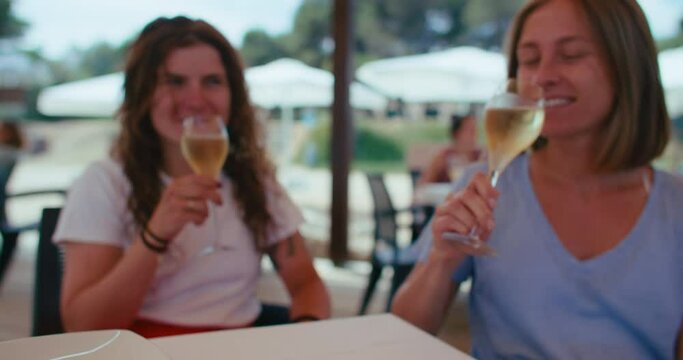Women couple celebrates their date, cheering and rising glasses with white sparkling wine, smiling and laughing on a terrace of a little beach bar
