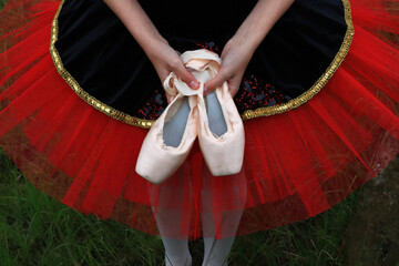 Looking down from above at the waist of a ballerina wearing a red and black tutu with gold colored trim holding her pink ballet pointe shoes in both hands. © Janice