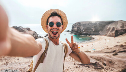 Handsome man wearing hat and sunglasses taking selfie picture on summer vacation day - Happy hiker with backpack smiling at camera outside - Tourist walking on the beach - Traveling and technology - obrazy, fototapety, plakaty