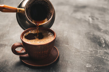 Pouring black coffee drink from turkish jezve coffee pot into clay cup on dark background