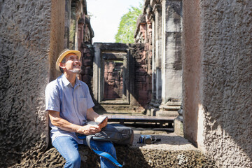 asian senior man traveller smiling and sitting on walkway the door of old architecture in Phimai...