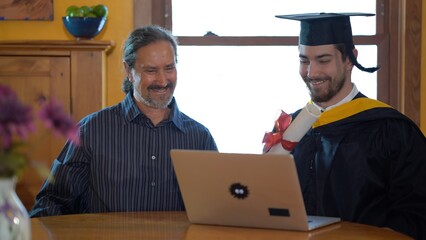Proud happy father and happy son sitting at dining room table doing online graduation ceremony...