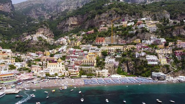 Aerial drone footage establish scene of Marina Grande beach, church of Santa Maria Assunta in Positano, Amalfi coast, Campania, Italy. Parasol, sun loungers, people swimming and colourful villas above