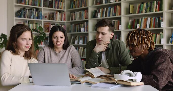 Multiethnic group of teen gen Z students watching virtual class, online learning presentation in library, attending conference, webinar, talking, discussing video at laptop and open books