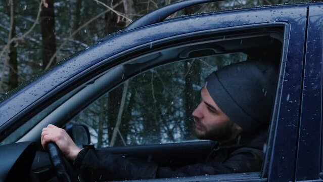 Young Caucasian Man Sits Behind Wheel Of Car With Open Window In Winter In Forest And Listens To Music, Dancing And Singing. Guy Driver Shakes His Head And Sings. Side View From Outside.