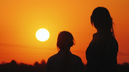 Mom and daughter look at the beautiful sunset over the city and the orange sky together