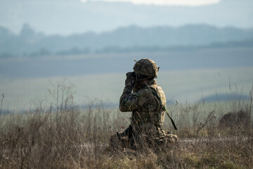 British army infantry soldier using binoculars to scan distance for enemy threats, military exercise, Wiltshire UK