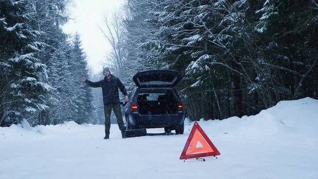Man got into trouble. Car broke down in winter on rural road during snowfall and guy driver standing and waiting for help, emergency stop sign red triangle to warn other drivers in foreground.