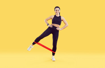 Fototapeta na wymiar Fitness woman standing and stretching elastic band with legs isolated on colored background. Full length woman with sporty body dressed in black leggings and sport top training on yellow background.