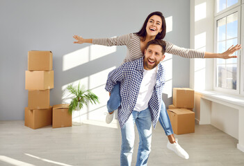 Cheerful family couple having fun on moving day. Happy, joyful young man piggybacks his wife or in...
