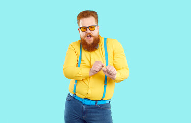 Funny cool stylish bearded fat man dancing victory dance isolated on light blue background. Caucasian red-haired millennial chubby man in jeans, shirt, suspenders and sunglasses dances waving his arms