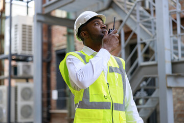 Fototapeta na wymiar African american male controller in protective hardhat using walkie talkie for regulation work and building process. Concept of safety engineer work at construction site.