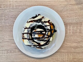 A big delicious cinnamon roll topped with white and dark chocolate sauce on a white plate on a wooden table. High quality photo