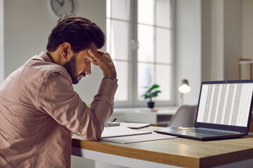 Tired, stressed business man sitting at his working desk with a laptop computer in the office, suffering from a headache and holding his hand on his head. Concept of stress, overworking and burnout