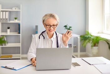 Professional doctor giving medical consultation. Happy woman in white coat with stethoscope sitting at office desk, video calling patient and waving hello at laptop computer. Online medicine concept - obrazy, fototapety, plakaty