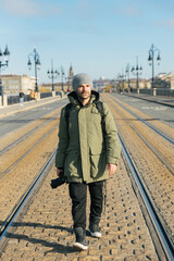 Young man with winter coat and knit holding photo camera walking through the middle of a bridge over train rails