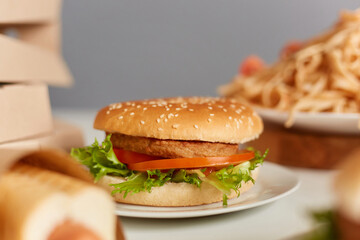 Closeup photo of hamburger with tomato on plate on table, hot dog and pasta on background, takeaway lunch, fast food snack.