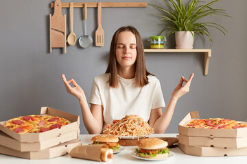 Portrait of calm relaxed grateful woman with brown hair wearing white casual T-shirt sitting at table in kitchen, practicing yoga or meditating before eating tasty dinner with pizza, pasta, hamburger. - obrazy, fototapety, plakaty