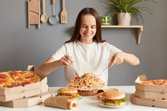 Image of beautiful optimistic woman with brown hair wearing white casual T-shirt sitting at table in kitchen, eating pasta with forks, smiling with satisfied face, enjoying junk food.