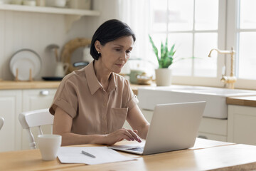 Focused senior freelance professional woman working from home, using laptop at kitchen table, typing, writing article. Serious mature 50s customer using online application for payment on Internet