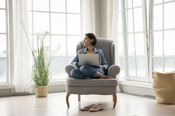 Thoughtful pretty middle aged lady relaxing with laptop in armchair, looking at window away, dreaming, enjoying leisure, calmness. Freelance business woman thinking on project, using computer at home