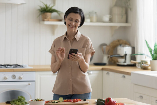 Cheerful happy mature chef woman using mobile phone over table with organic food ingredients, cooking salad, reading recipe on Internet, taking picture, smiling, laughing - Powered by Adobe