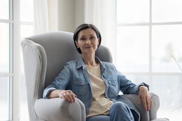 Positive senior 50s woman sitting in armchair, looking at camera, smiling. Pretty brunette mature...