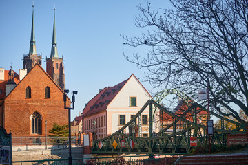Obraz premium Two tall towers under a metal roof with crosses behind three houses. A bridge across the Oder in the foreground. Cityscape Wroclaw Poland, Tumsky Island.